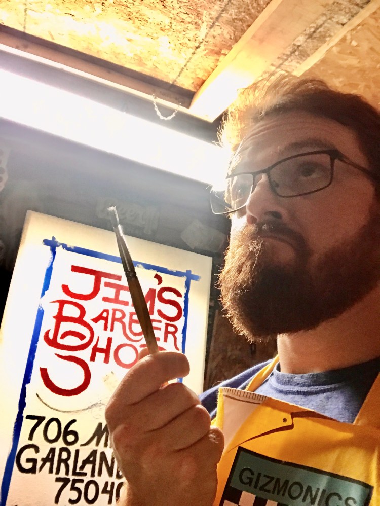 Photo of Will in his workshop, holding a paintbrush in front of a sign he's painting with partially obscured text on it: "Jim's Barber Shop, 706 M, Garland 75040".