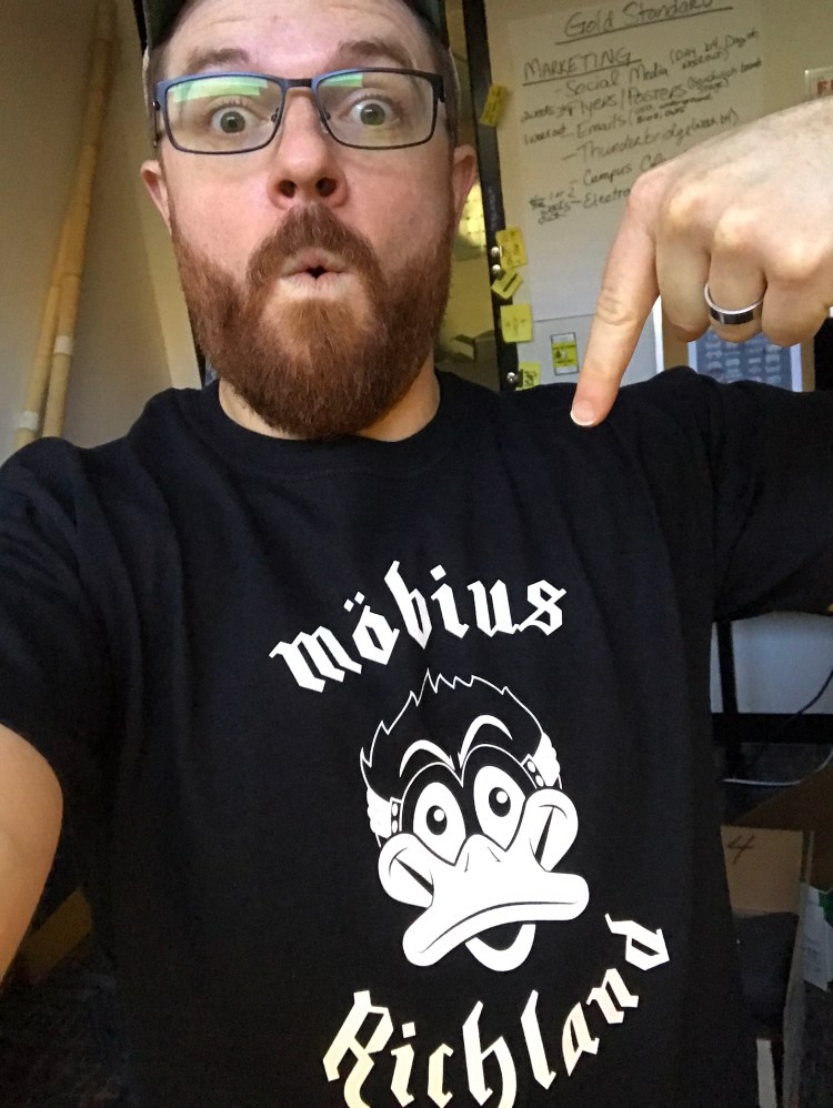 A photo of Will pointing at his own shirt he designed after the famous Mötorhead shirt. The shirt features a white image of Möbius on black with the text "Möbius Richland".